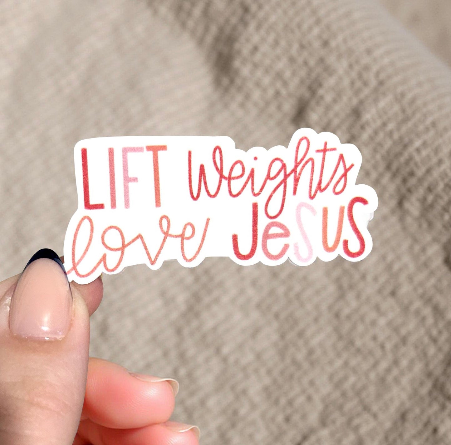 Hand holding a sticker with 'LIFT Weights love Jesus' text against a beige background