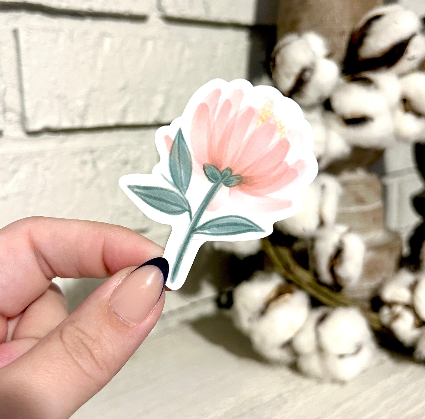 Hand holding a peony sticker with a textured wall and decorative item in the background