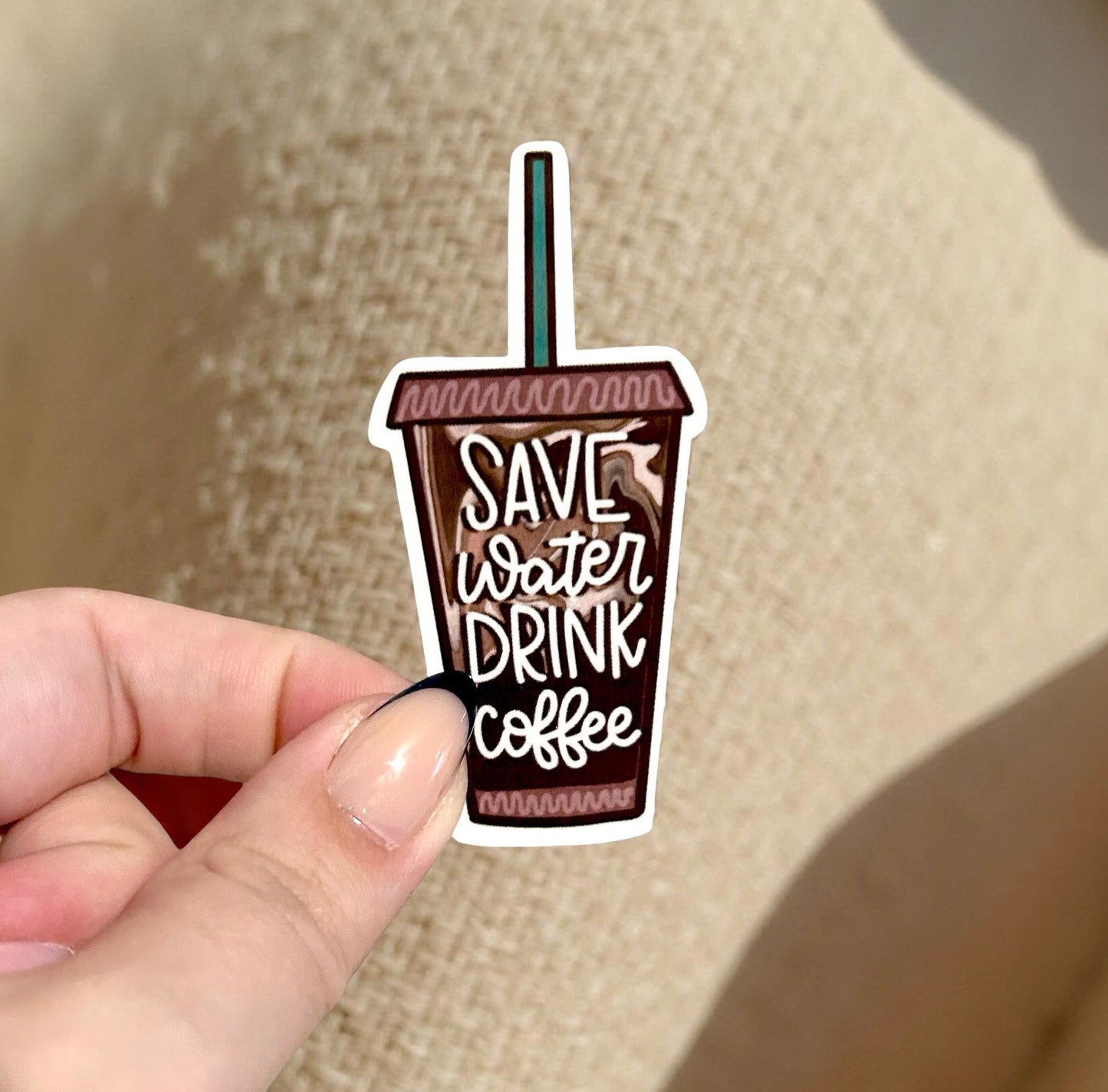 Sticker shaped like a coffee cup with 'Save water, drink coffee' text held by a hand on a beige background.