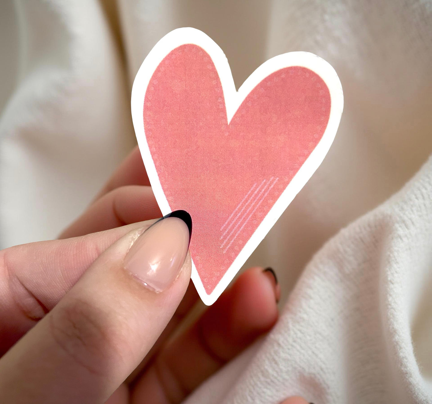 Heart-shaped pink sticker held by a hand against a neutral background
