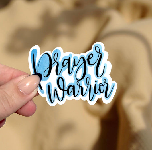 Hand holding a 'Prayer Warrior' sticker against a blurred background