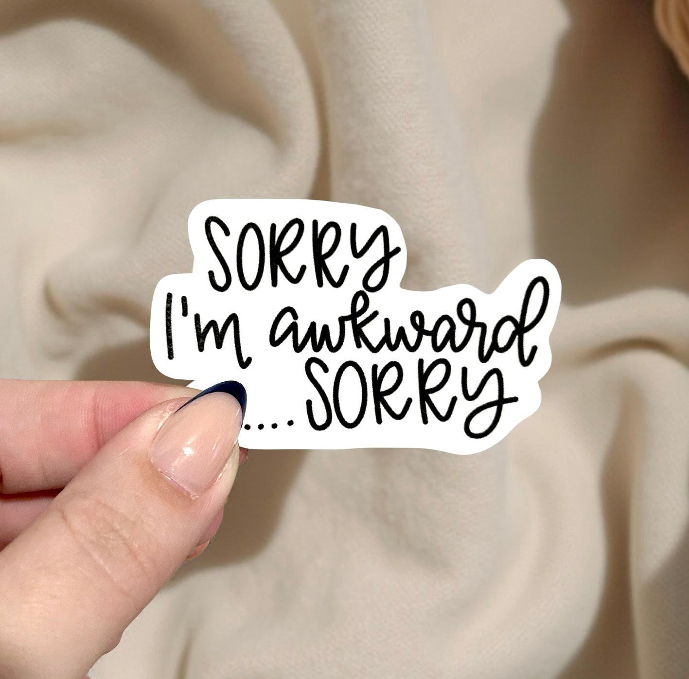 Hand holding a sticker with 'Sorry I'm awkward... Sorry' text against a beige fabric background