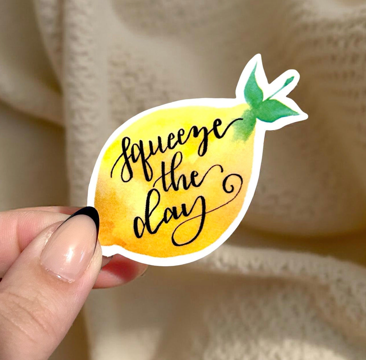 Hand holding a lemon-shaped sticker with 'squeeze the day' text against a textured beige background.