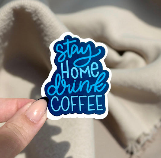 Sticker with 'Stay Home Drink Coffee' text held by a hand against a textured beige background