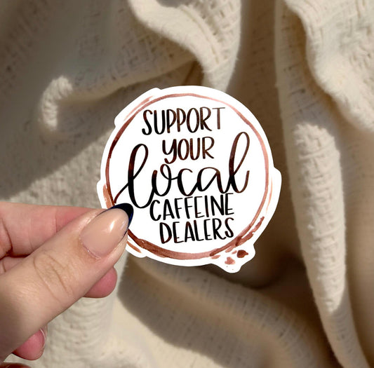 Hand holding a sticker with 'Support your local caffeine dealers' text against a textured beige background.