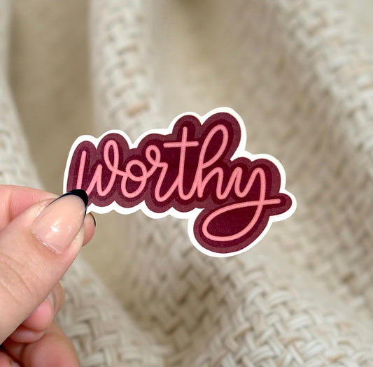 Hand holding a 'Worthy' sticker against a textured beige background