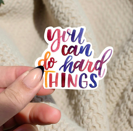 Hand holding a colorful sticker with 'you can do hard things' text against a textured beige background.