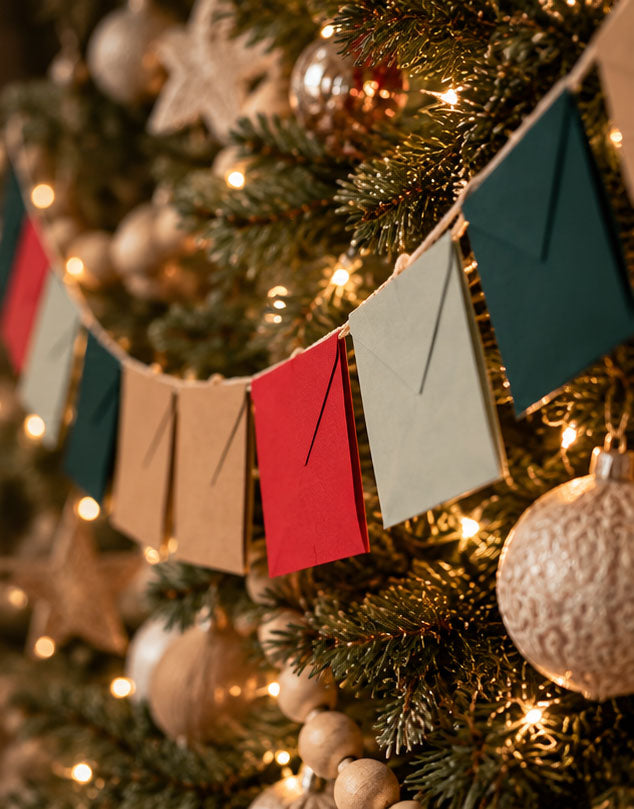 A festive Christmas tree decorated with warm white string lights, gold ornaments, and a hanging paper banner made of square flags in red, beige, light gray, and dark teal.