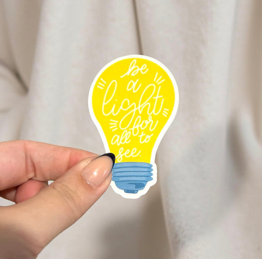 Hand holding a yellow light bulb sticker with blue base and text 'Be a light for all to see' against a beige background.