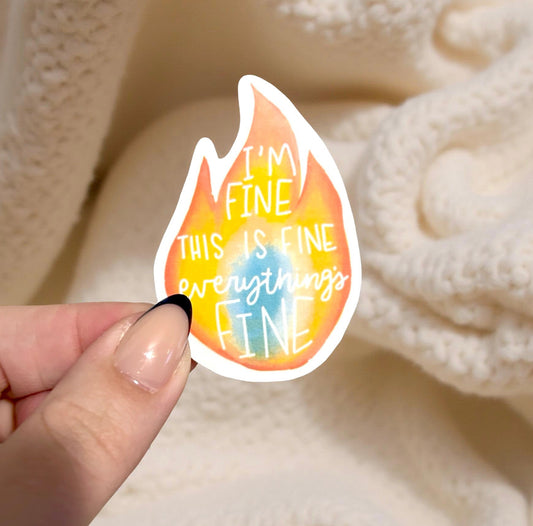 Hand holding a colorful sticker with text 'I'm fine, this is fine, everything is fine' against a soft beige background.