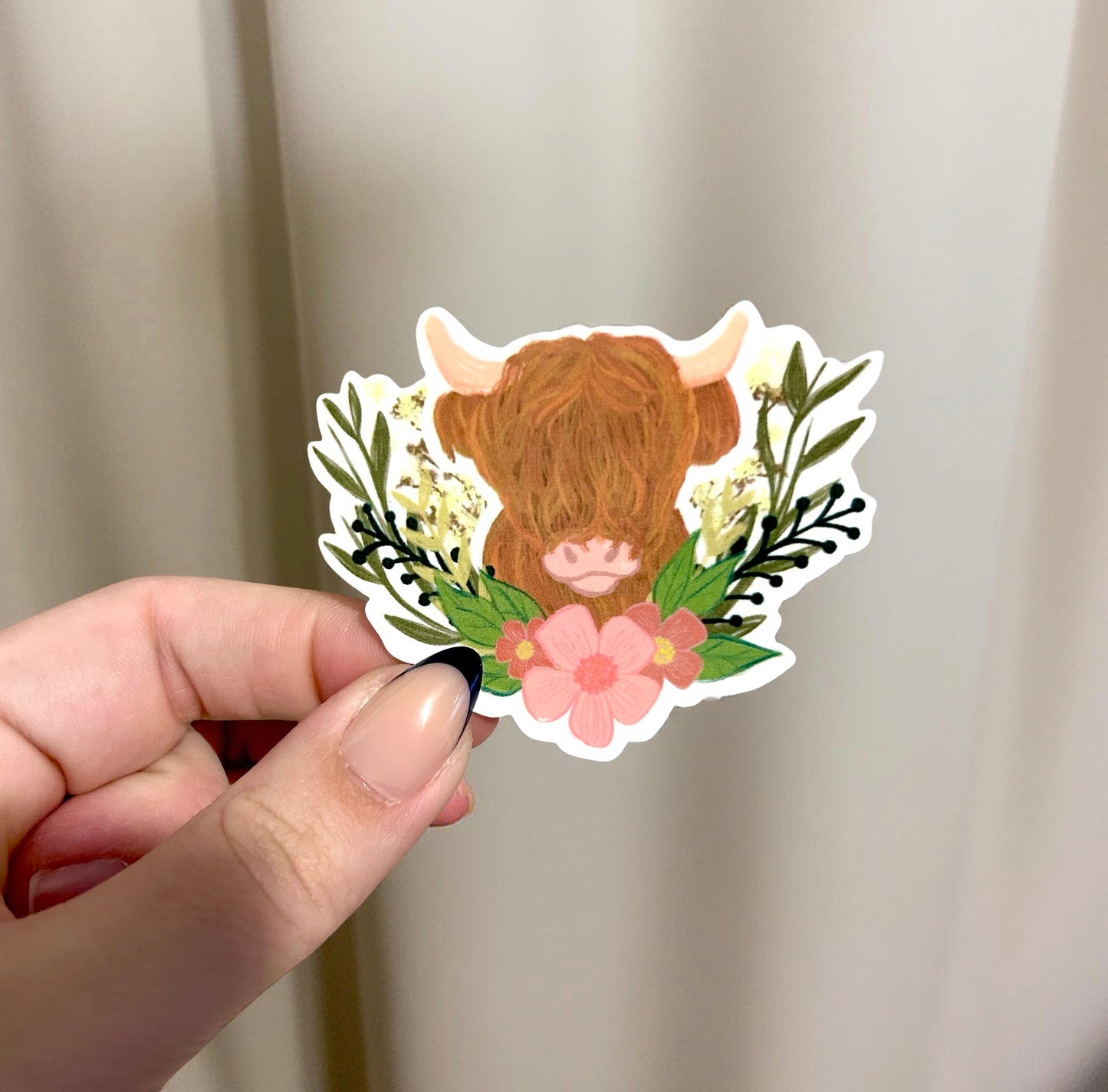 Hand holding a sticker of a cow surrounded by flowers and leaves against a plain background