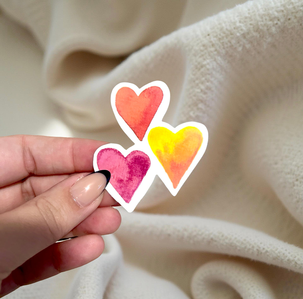 Hand holding three colorful heart-shaped stickers against a beige fabric background